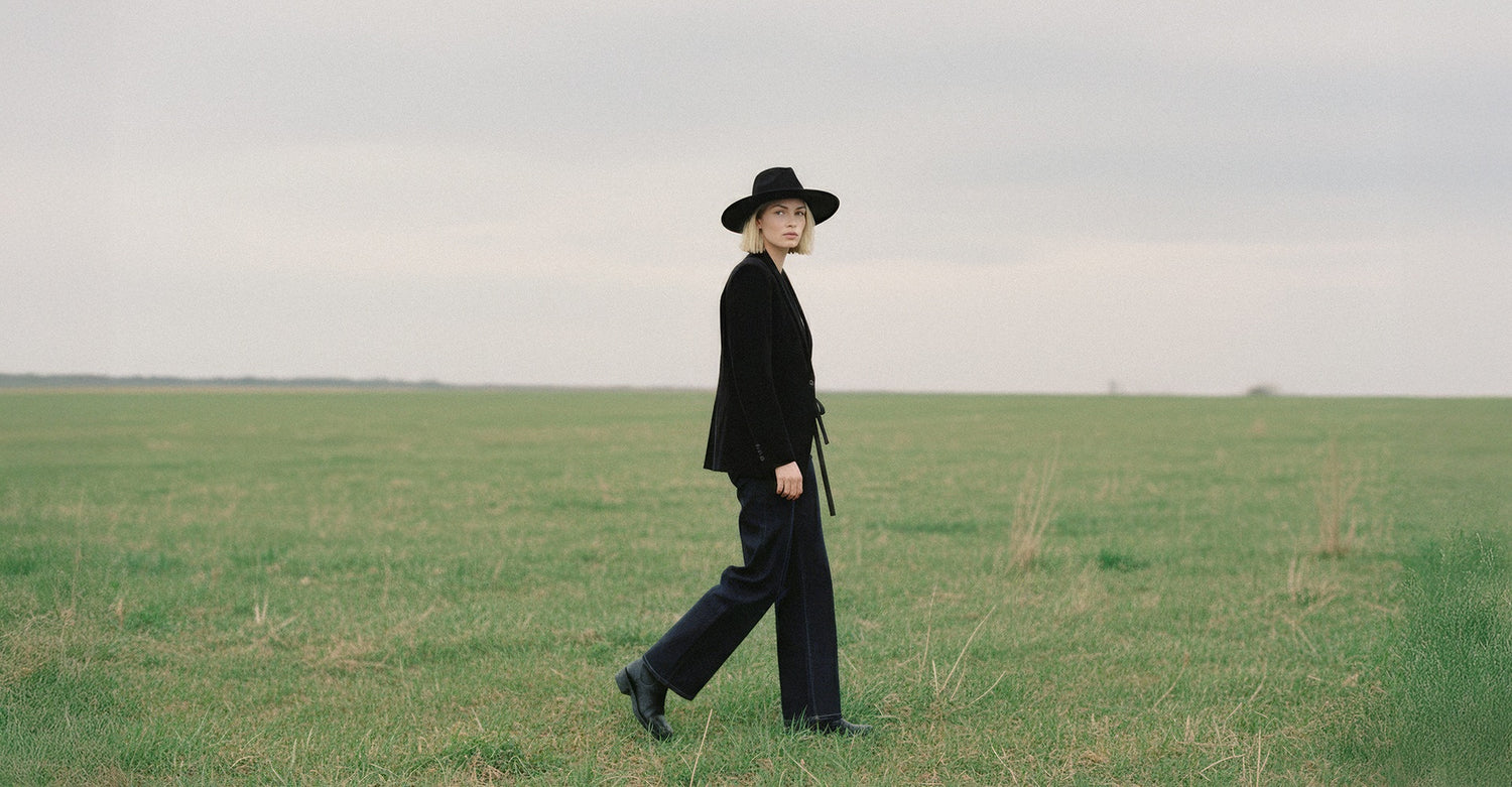 Model walking outdoors through an open grass field wearing dark wash wide-leg denim jeans, a black jacket, and a wide-brim hat.